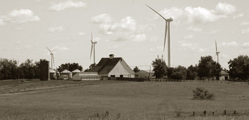 A rural farm in the foreground with four wind turbines in the background.